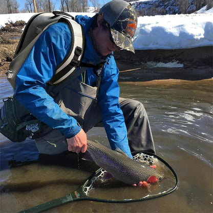 Person carry with Buffalo Gear Fishing Backpack in blue jacket and gray pants holding a fish in a snowy landscape with a river.